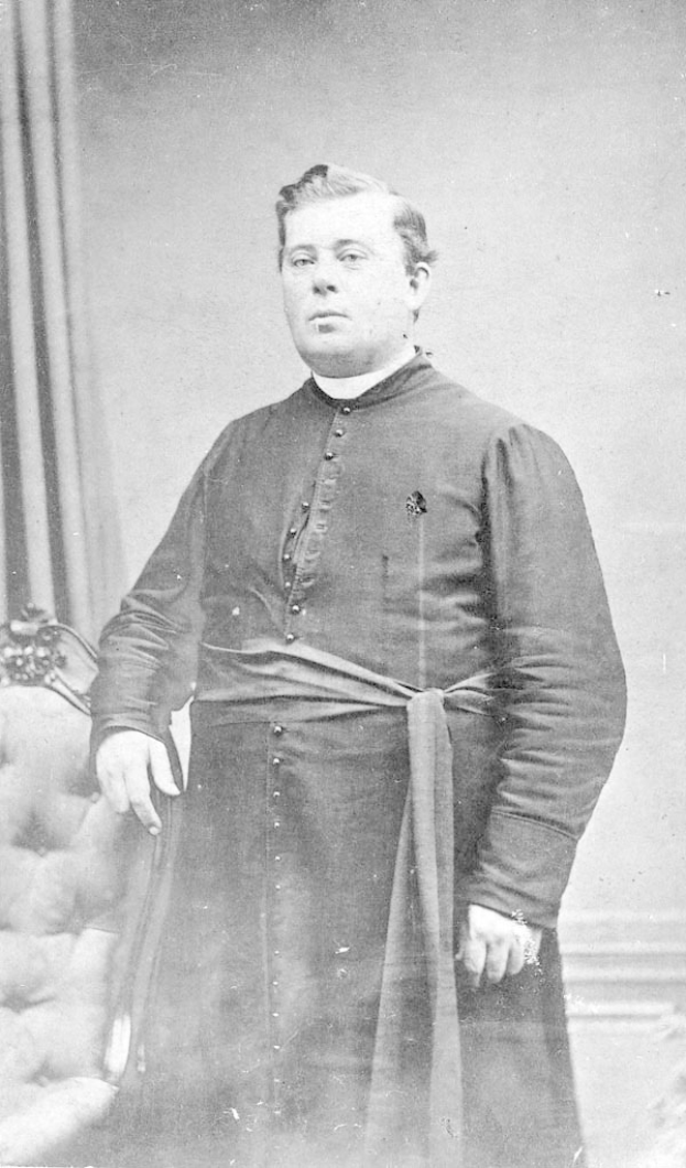 Schwarzes und weißes Foto eines Mannes in Priesterroben, identifiziert als John F. Kennedy, erster Bischof der Kirche von England, der vor einer Vorhangwand steht.