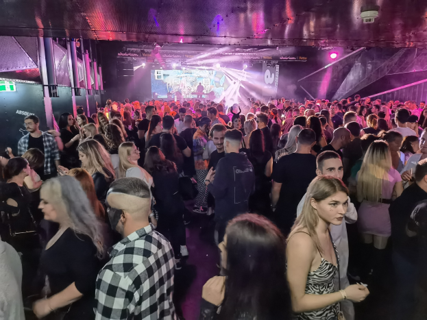 Eine große Menschenmenge in einer Diskothek mit Deckenbeleuchtung und Säulen, mit einem Bildschirm im Hintergrund, der auf ein Absolut Nightlife-Event hinweist.