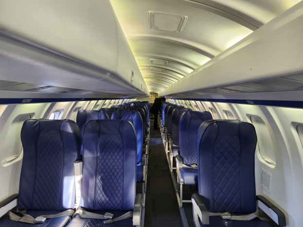 Leere Flugzeugsitze mit einer Person im Gang, bezogen mit hellen Stoffen, große Fenster und Deckenbeleuchtung.