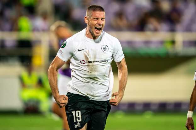 Ein Mann in weißem Hemd und schwarzen Shorts läuft auf einem Fußballfeld, feiert ein Tor, umgeben von einer Gruppe von Menschen, mit unscharfem Hintergrund.