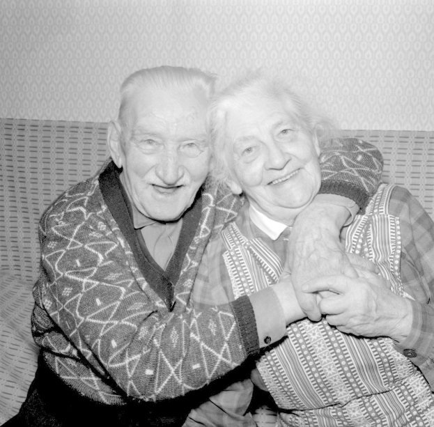 Schwarzes und weißes Foto eines älteren Paares auf einem Sofa sitzend, das sich umarmt und lächelt.