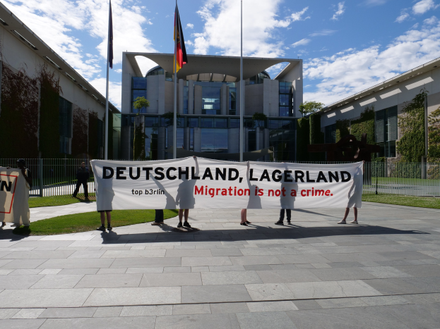 Eine Gruppe von Menschen hält ein Transparent mit der Aufschrift 'Deutschland, Lagerland Migration ist kein Verbrechen' vor einem Gebäude mit Fenstern, umgeben von Gras, einem Metallzaun, Pflanzen, Bäumen und Fahnen auf Stangen unter einem bewölkten Himmel.