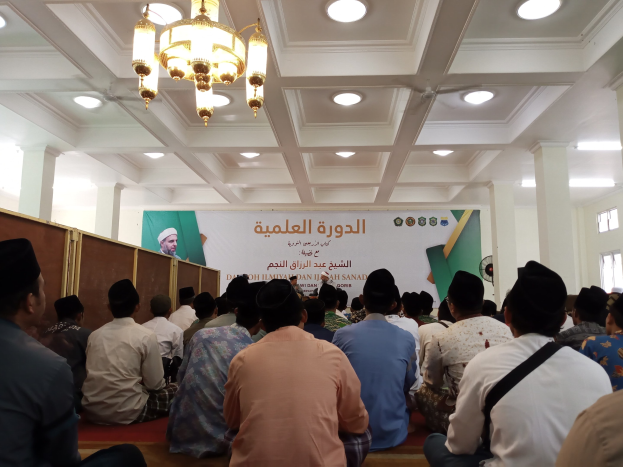 Gruppe von Menschen, die auf dem Boden in einem hellen Saal mit Säulen, einem Banner mit Text und einem Personenbild sowie einem Fenster auf der rechten Seite sitzen und an einem Seminar zum Islam teilnehmen.