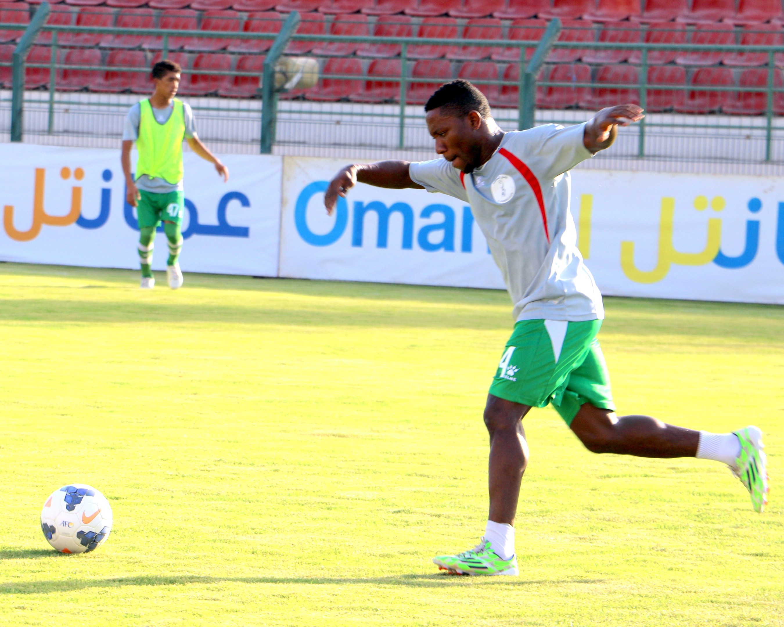Ein Mann tritt einen Fußball auf einem Rasenfeld in einem Stadion, mit zwei Personen in der Nähe und Bannern, Geländern und Stühlen im Hintergrund.