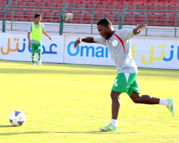 Ein Mann tritt einen Fußball auf einem Rasenfeld in einem Stadion, mit zwei Personen in der Nähe und Bannern, Geländern und Stühlen im Hintergrund.
