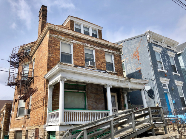 Eine Reihe von baufälligen Häusern mit kaputten Fenstern, bröckelnden Wänden, Geländern, Stufen, einem Zaun, einer Satellitenschüssel und Überlandleitungen, vor einem bewölkten Himmel.