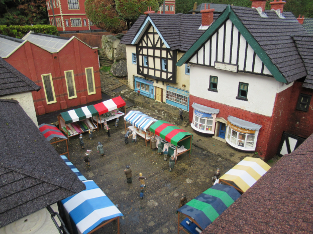Ein detailliertes Miniaturmodell einer Kleinstadt mit Gebäuden, Häusern, Zelten, Menschen, Bäumen, Pflanzen, Felsen, einer Straße unten und einem zentralen Marktstand.