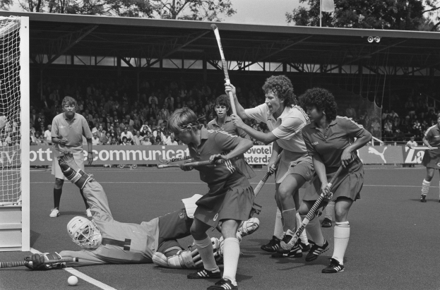Eine Gruppe von Frauen, die Feldhockey spielen, in Schwarz-Weiß, mit Helmen und Hockeystöcken, einem Torpfosten links, Zuschauern auf den Tribünen, Bannern, einem Schuppen, Bäumen und einer Fahne im Hintergrund.