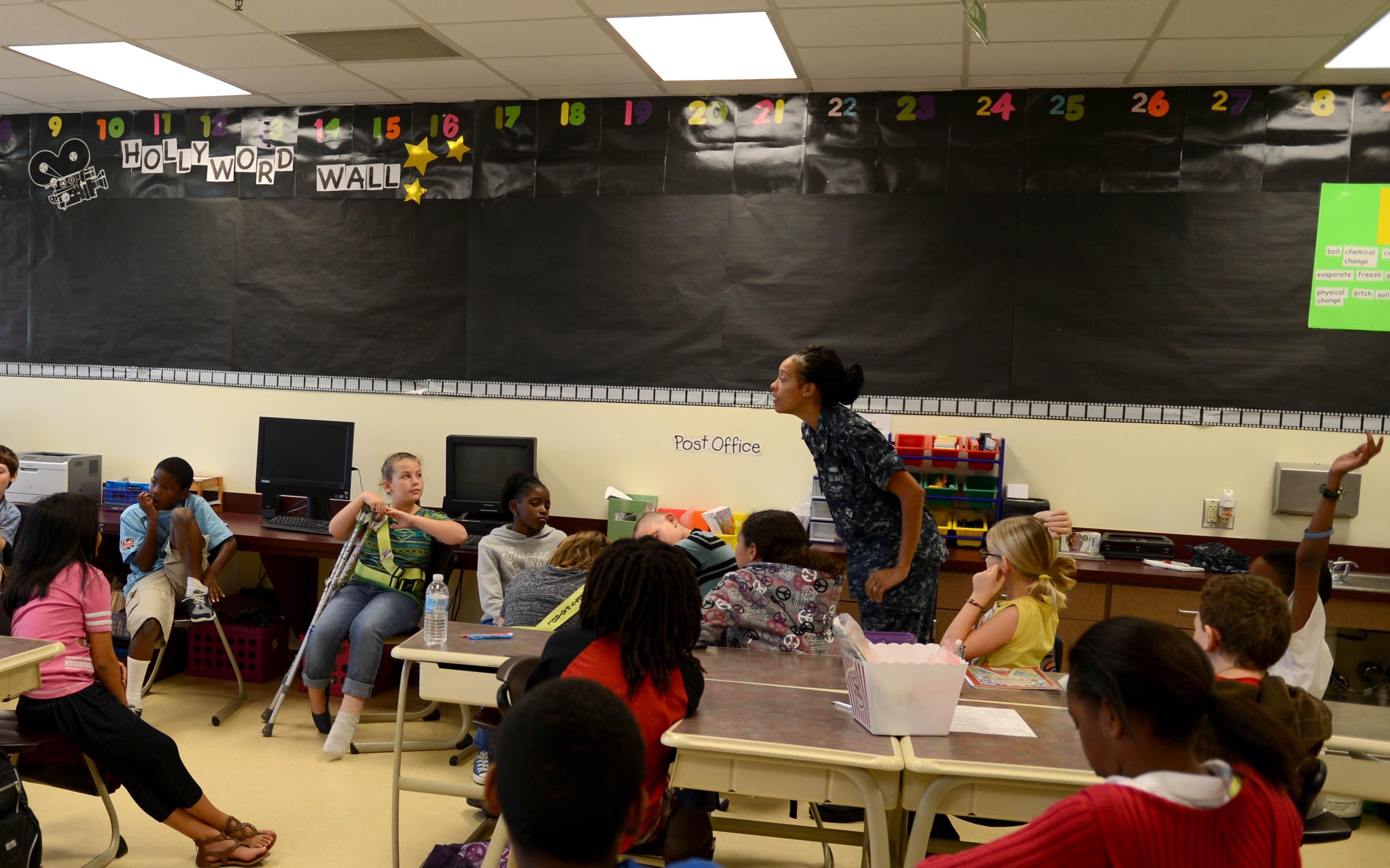 Ein Klassenzimmer mit Kindern an Tischen mit verschiedenen Gegenständen, einem Lehrer in der Mitte und einer Tafel im Hintergrund.