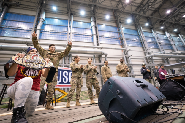 Eine Gruppe von Menschen auf einer Bühne mit einer Frau, die in der Hocke ist und einen Gürtel hält, Lautsprecher und Equipment auf dem Boden und eine Tafel im Hintergrund mit der Aufschrift "WWE World Heavyweight Championship: The Best of the Best."
