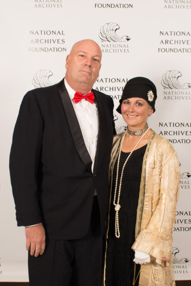 Ein Mann in einem schwarzen Anzug mit weißem Hemd und schwarzer Fliege steht neben einer Frau in einem eleganten Kleid mit einem gemusterten Schal auf einem roten Teppich, mit einem weißen Banner im Hintergrund.