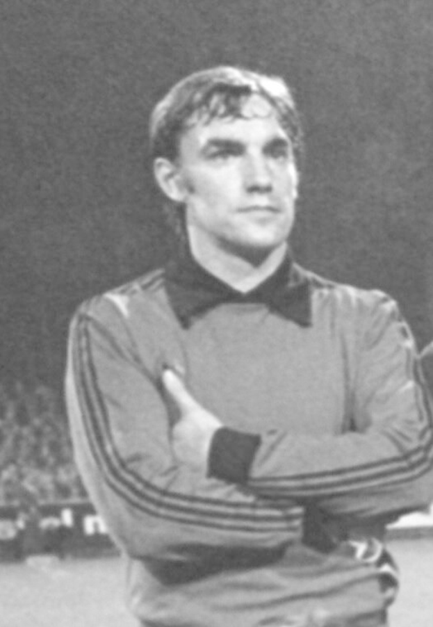 Black and white photo of a man standing on a soccer field with arms crossed, surrounded by a group of seated people in the background.