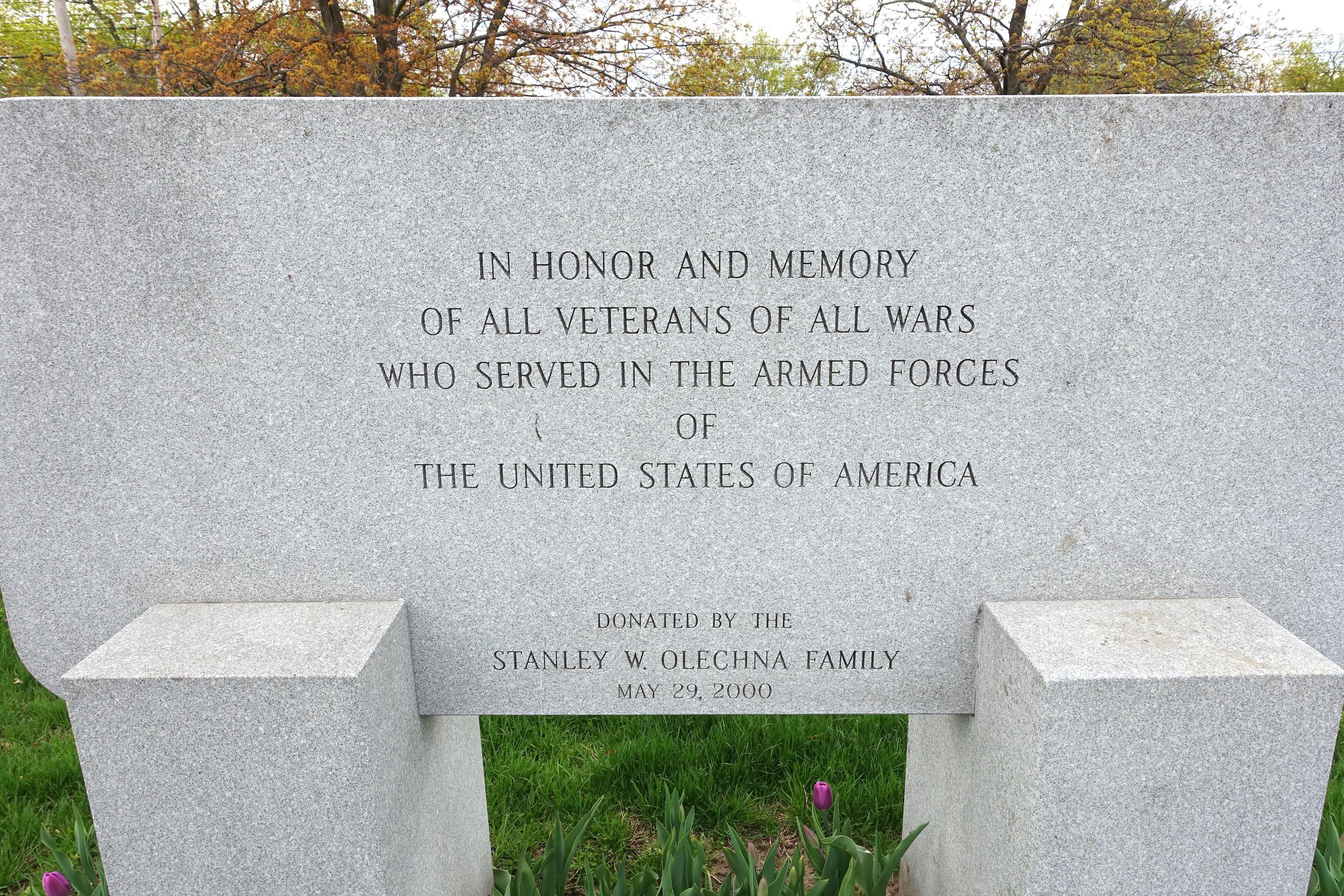 Denkmalstein mit der Inschrift 'In Ehren und Andenken an alle Veteranen aller Kriege, die in den Streitkräften der Vereinigten Staaten von Amerika gedient haben', umgeben von Pflanzen, Blumen, Gras, Bäumen und einem bewölkten Himmel.