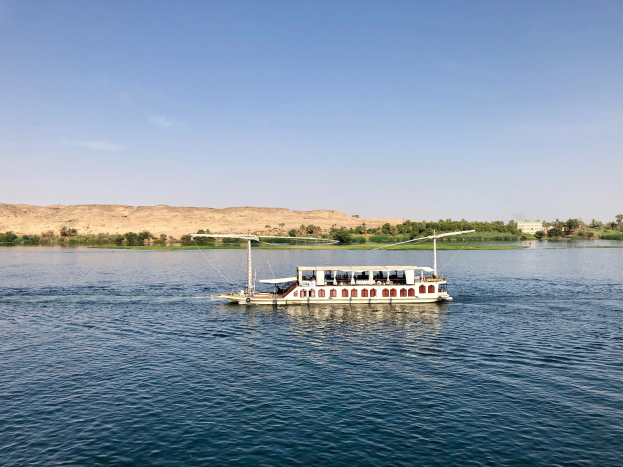 Ein Boot fährt den Nil in Ägypten hinunter, umgeben von grünen Bäumen unter einem hellblauen und weißen Himmel, wahrscheinlich Teil einer Nilkreuzfahrt.