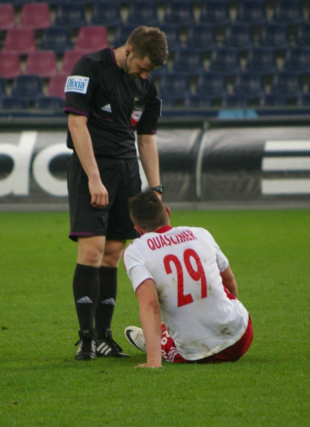 Ein Fußballspieler und Schiedsrichter sitzen auf dem Boden in einem Stadion, beide in Sportkleidung gekleidet, mit Brettern und Stühlen im Hintergrund.