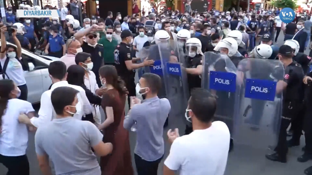 Eine Gruppe von Menschen vor einer Reihe von Polizei-offizieren mit Helmen und Schilden, mit einem geparkten Auto links und einer Person mit einer Kamera rechts, vor Gebäuden und Bäumen in Ankara, Türkei.