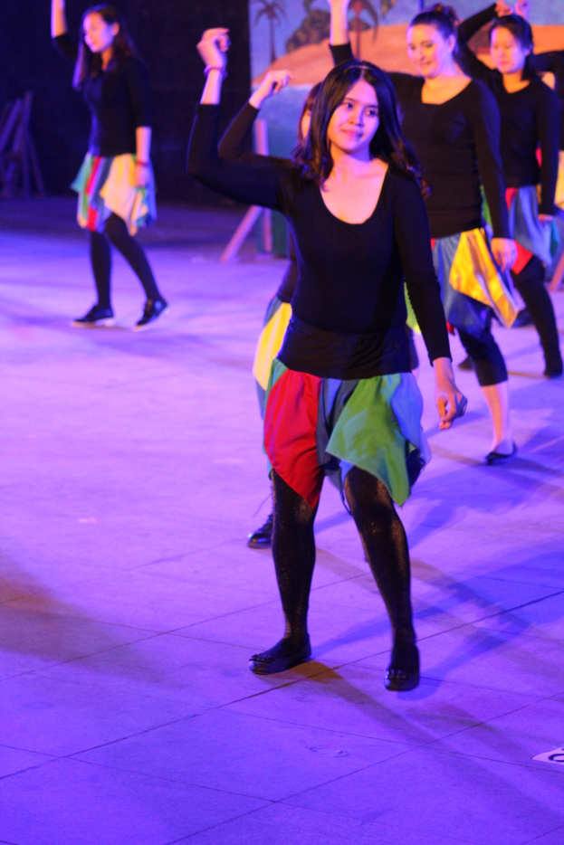 Gruppe junger Frauen, die auf einer Bühne tanzen, mit einem Gemälde an der Wand dahinter, alle tragen Schuhe während einer Vorstellung.