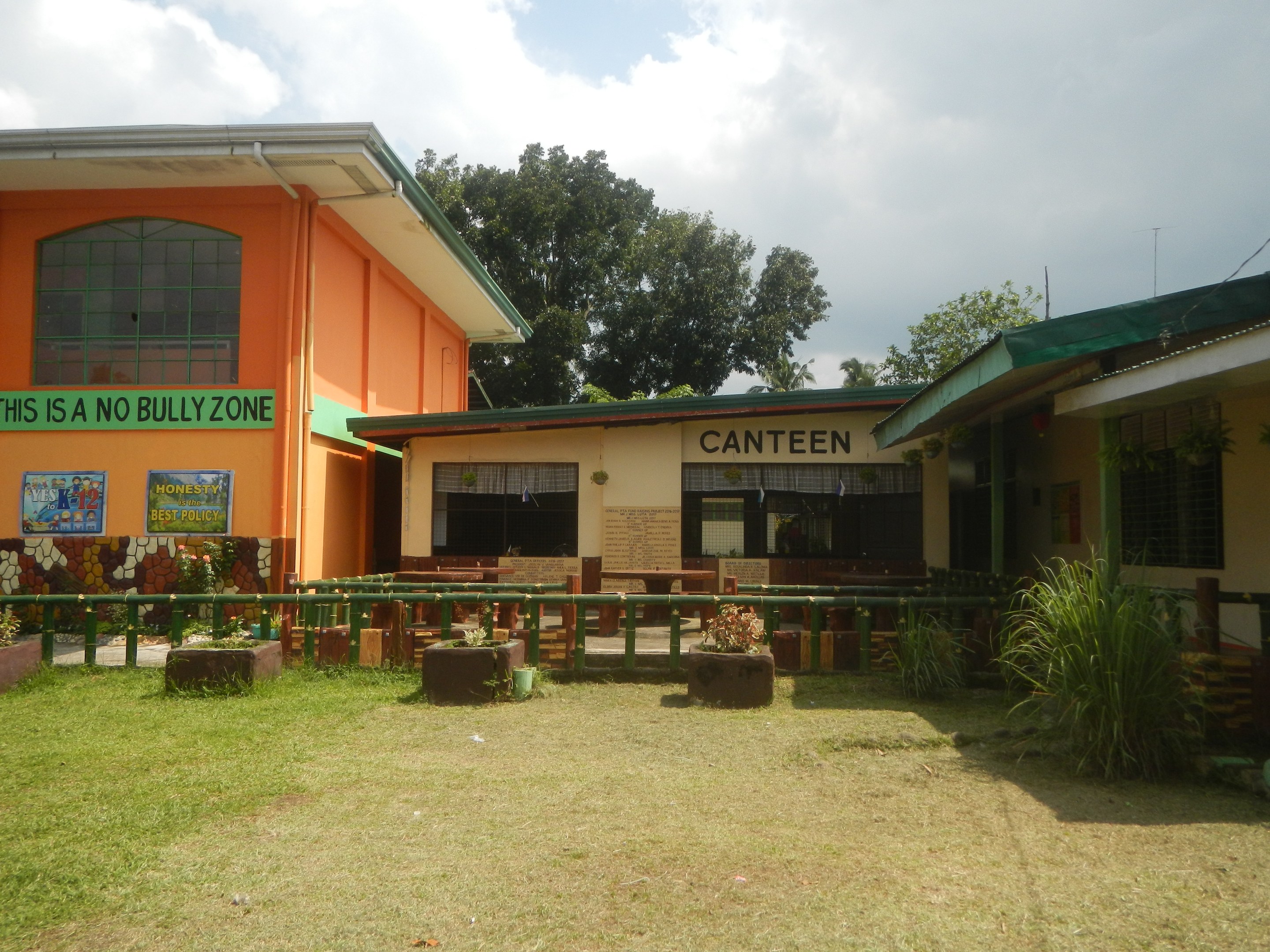 A building with a "This is a No Bullying Zone" sign, surrounded by houses, greenery, a fence, trees, and a cloudy sky.