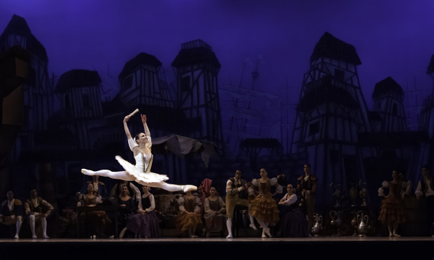 Eine Ballerina in einem weißen Tutu und Ballettschuhen tanzt auf der Bühne, umgeben von einem Publikum, das sitzt und steht, und einem Hintergrundbild eines Schlosses.