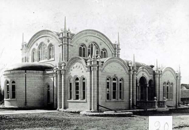 Ein Schwarz-Weiß-Foto eines großen Gebäudes mit Bögen, Säulen und Fenstern, umgeben von Bäumen und einem klaren Himmel, mit Text unten, der es als Postamt identifiziert.