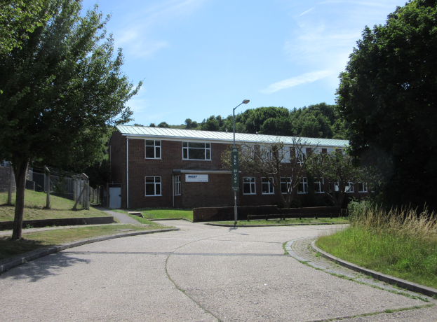 Ein Schulgebäude mit Fenstern, einem Straßenschild, einer Tafel, einer Gruppe von Bäumen, Gras, einem Zaun und einem bewölkten Himmel, das in der Mitte eines Parks umgeben von saftigem Grün liegt.