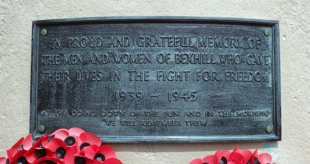 Eine Metallgedenktafel an der Außenseite eines Gebäudes mit eingraviertem Text, begleitet von roten Mohnblumen davor, die Leben im Dienst verloren haben.