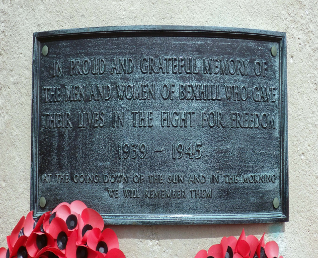 Eine Metallgedenktafel an der Außenseite eines Gebäudes mit eingraviertem Text, begleitet von roten Mohnblumen davor, die Leben im Dienst verloren haben.