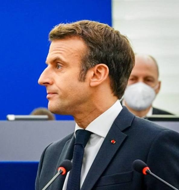 Ein Mann im Anzug und Krawatte steht an einem Podium mit zwei Mikrofonen, während eine maskierte Person hinter ihm steht, mit einem unscharfen Hintergrund.