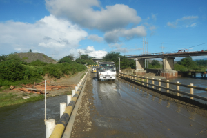 Ein Laster fährt auf einer überfluteten Straße neben einer Brücke mit Geländern, mit Bäumen, Pfählen und einem bewölkten Himmel im Hintergrund.