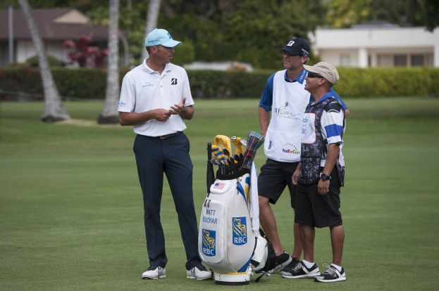 Gruppe von Männern auf einem grünen Feld mit einem Golfbag und -schlägern im Vordergrund, Bäume, Pflanzen und Gebäude im Hintergrund.
