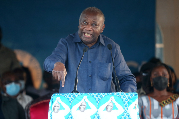 Ein Mann in einem blauen Hemd spricht an einem Podium mit zwei Mikrofonen vor einer sitzenden Menge, wobei einige Masken tragen, und eine Wand im Hintergrund zu sehen ist.