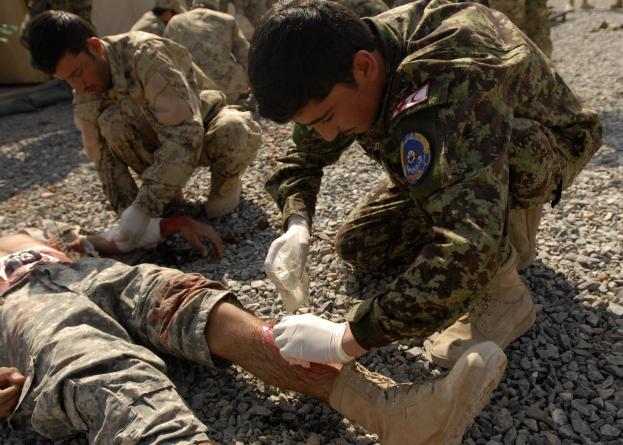 Eine Gruppe von Männern in Militäruniformen kniet auf dem Boden, einer liegt mit einem verbundenen Bein, während die anderen im Hintergrund bei einem Zelt stehen.