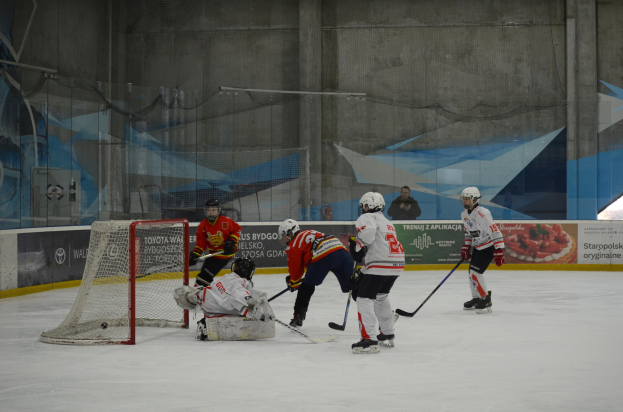 Gruppe von Menschen, die Eis hockey auf einem Eisplatz spielen, tragen Helme und halten Hockey-Schläger, mit einem Tor auf der linken Seite und Bannern im Hintergrund.