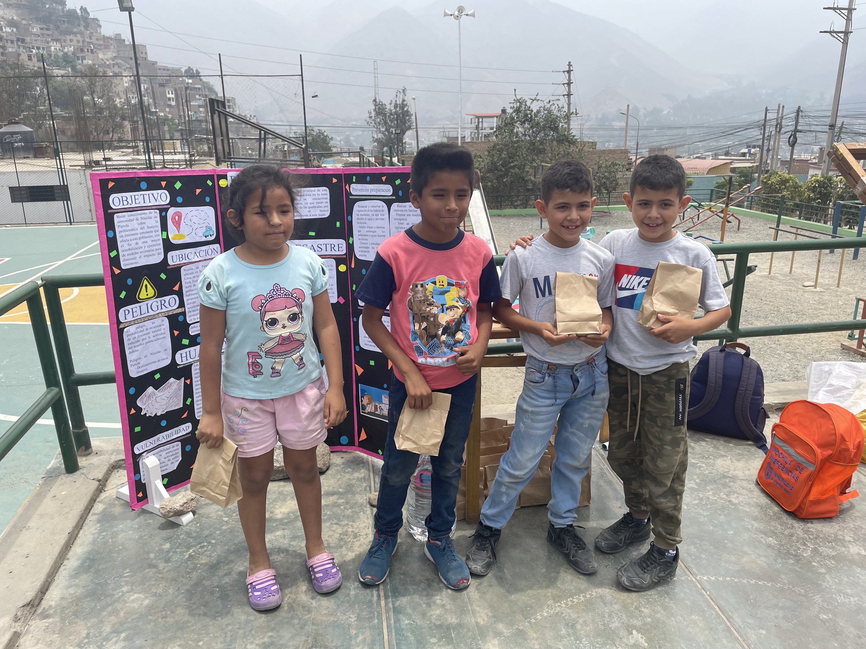 Gruppe von Kindern, die Papiertüten auf einem Basketballfeld halten, mit Tüten und Gegenständen auf dem Boden, einer Tafel mit Text links daneben und einem Hintergrund mit Zaun, Pfosten, Straßenlaternen, Bäumen, Gebäuden, Hügeln und Himmel.