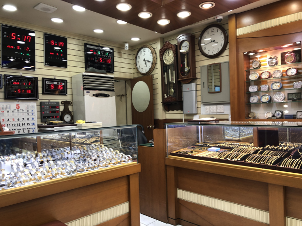 Ein Schmuckgeschäftsinneres mit Glasvitrinen, die mit Uhren gefüllt sind, flankiert von Bildschirmen und einer Wand mit Uhren im Hintergrund, beleuchtet von Deckenlampen.