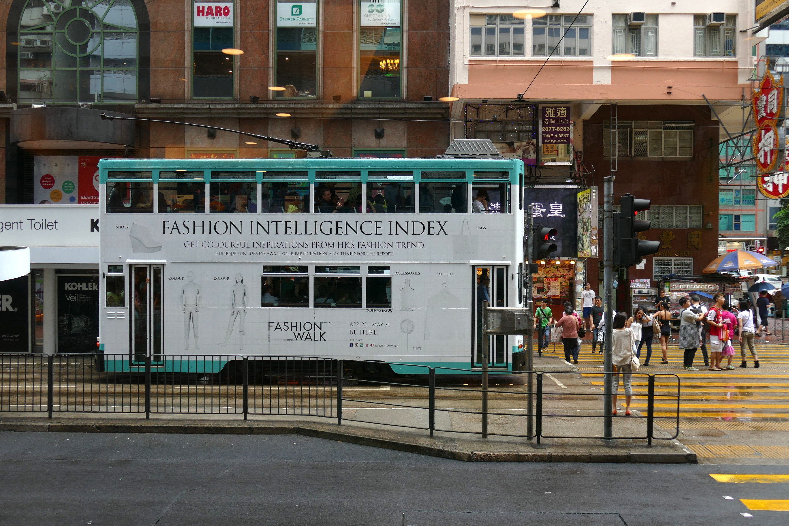 Doppeldeckerbus fährt auf einer städtischen Straße mit hohen Gebäuden, Passanten auf dem Gehweg, einige halten Regenschirme, Verkehrsampeln, ein Geländer, Informationsschilder, Bäume und einen klaren blauen Himmel.