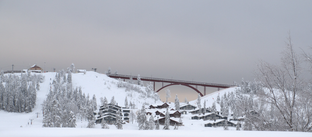 Ein Skiresort mit einer Brücke über einen schneebedeckten Hügel, schneebedeckte Bäume und Gebäude, einen klaren blauen Himmel und Strommasten mit Drähten im Hintergrund.