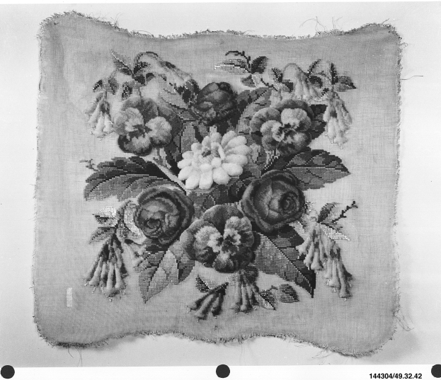 Schwarzes und weißes Foto eines Stoffbouquets mit genähten Blumen, grünen Blättern und Stielen, die in einer Vase auf einem Tuch angeordnet sind.