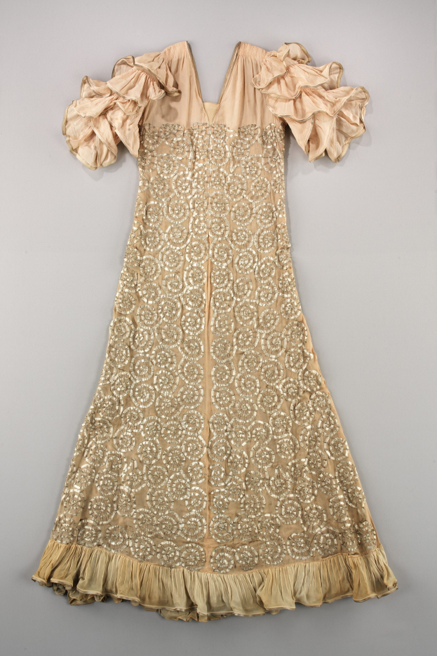 Ein cremefarbenes Kleid mit goldener Stickerei und Rüschen an den Ärmeln, das auf einer weißen Oberfläche präsentiert wird und detaillierte Muster und Designs aufweist.