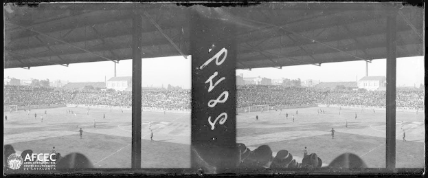 Schwarzes und wei├čes Foto eines Fussballspiels in einem Stadion mit Spielern auf dem Feld und Zuschauern in den R├Ąngen, vor Geb├Ąuden, B├Ąumen und einem klaren Himmel, mit dem Text und Logo "Piazza della Repubblica, Rom, Italien" unten links.