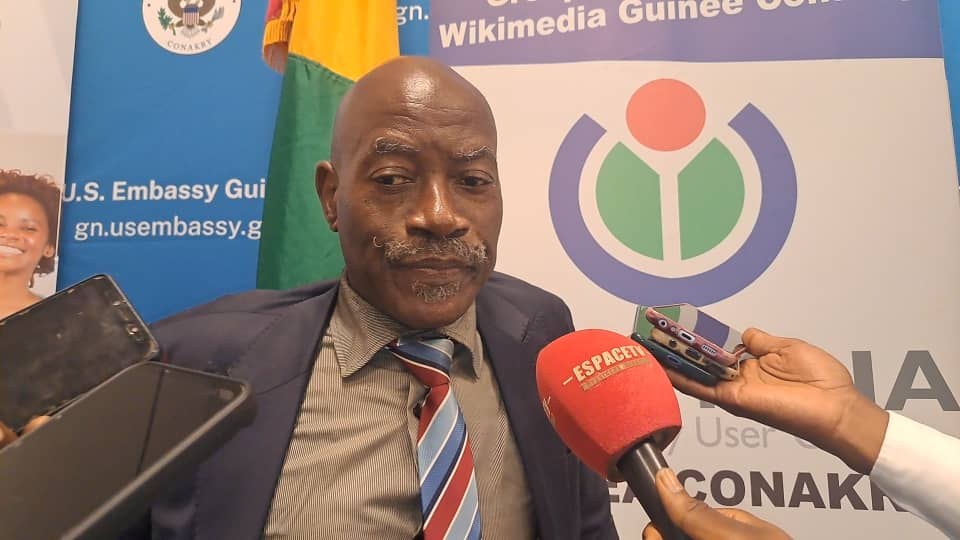 Ein Mann im Anzug spricht mit Reportern, Mikrofon und Aufzeichnungsgerät im Vordergrund, Banner, Flaggen und eine Frau im Hintergrund.