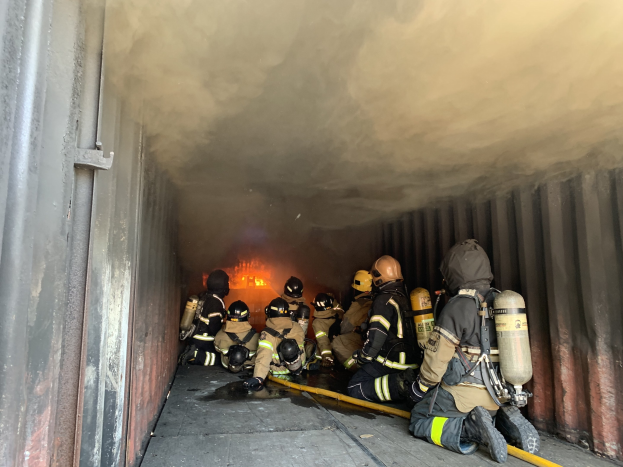 Feuerwehrleute in Schutzanzügen arbeiten in einem raucherfüllten Tunnel, einer hält einen Schlauch, mit Flammen im Hintergrund und einer Wand auf der linken Seite.