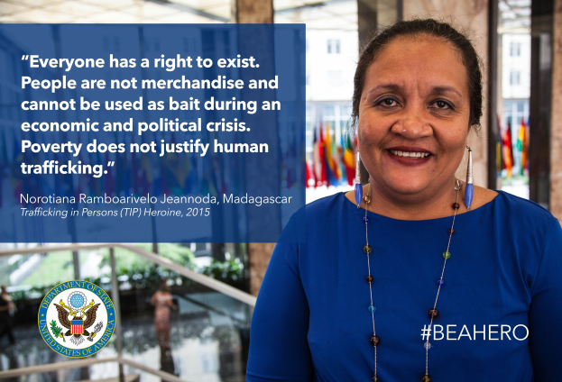 Eine Frau in einem blauen Kleid steht lächelnd vor einem Gebäude mit einer Inschrift über Menschenhandel, umgeben von Menschen, Pflanzen und Flaggen.