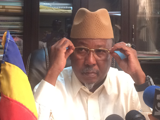 Ein Mann in einem weißen Hemd und Hut sitzt an einem Schreibtisch mit Mikrofonen, trägt eine Brille, eine Uhr und eine Flagge links von ihm und spricht in einer Bibliothek mit Bücherregalen hinter ihm.