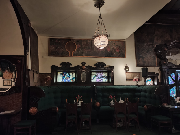 Innenraum eines gut eingerichteten Restaurants mit Tischen, Stühlen, Blumenvasen, Sofas, einem Kronleuchter, gerahmten Kunstwerken an den Wänden und einem Fenster auf der rechten Seite.