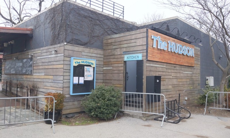 Außenansicht des Hudson-Restaurants in New York City, mit einer Tür, einem Schild, Topfpflanzen, einem Metallzaun, Bäumen und einem Mülleimer unter einem bewölkten Himmel.