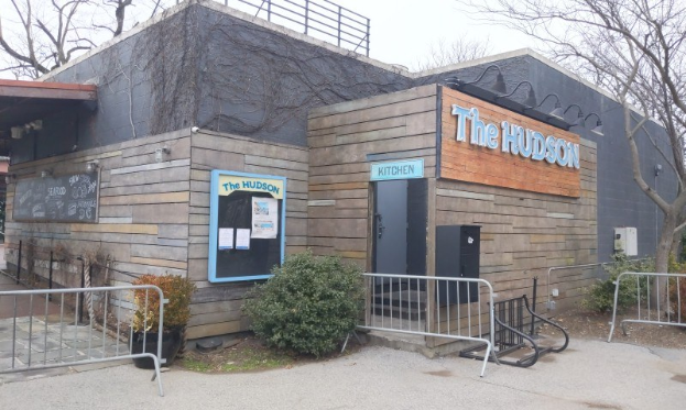 Außenansicht des Hudson-Restaurants in New York City, mit einer Tür, einem Schild, Topfpflanzen, einem Metallzaun, Bäumen und einem Mülleimer unter einem bewölkten Himmel.