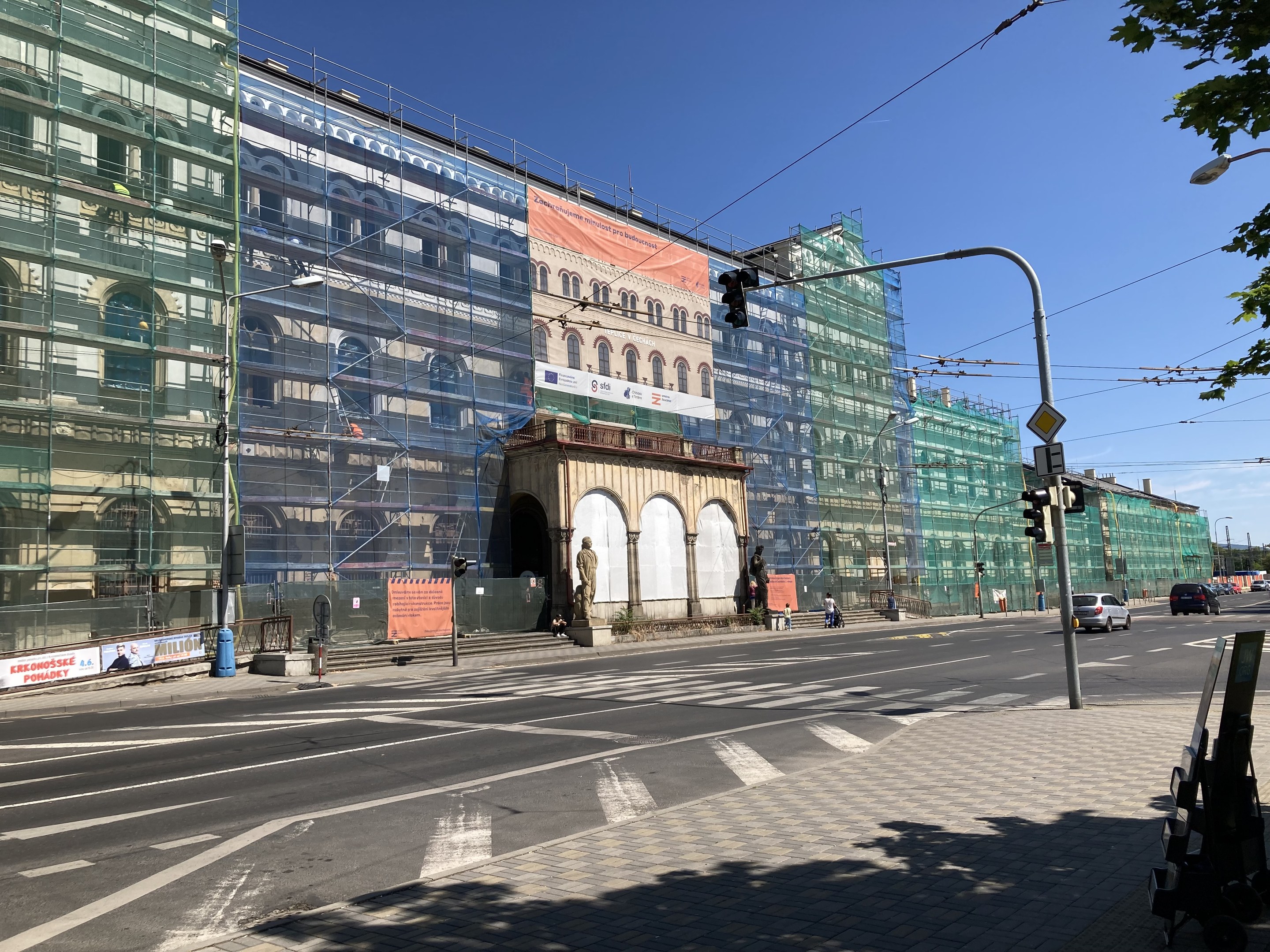 Großes mehrstöckiges Gebäude im Bau in Berlin mit Gerüst, umgeben von Straßeninfrastruktur, Fahrzeugen, Fußgängern und Bäumen bei einem bewölkten Himmel.