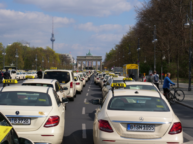 Eine lange Reihe von Taxis, die an einer belebten Straße in Berlin, Deutschland, geparkt sind, mit Fahrzeugen, Radfahrern und Fußgängern, flankiert von Laternenpfählen, Bäumen und Gebäuden, einschließlich eines Bogens und eines Turms im Hintergrund unter einem bewölkten Himmel.