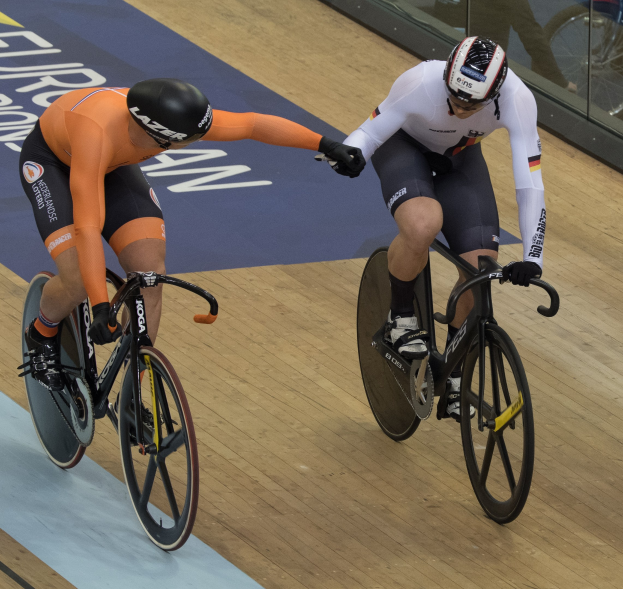 Zwei Radrennfahrer mit Helmen auf einer Velodrom-Bahn mit Glaswand im Hintergrund.
